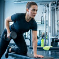 Une femme concentrée effectue un tirage haltère à un bras sur un banc de musculation dans une salle de sport.