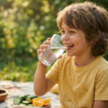 Jeune garçon souriant buvant un verre d'eau fraîche en extérieur lors d'un pique-nique ensoleillé.