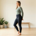 Une femme debout de profil dans une salle de sport lumineuse, portant un haut vert et un legging noir.